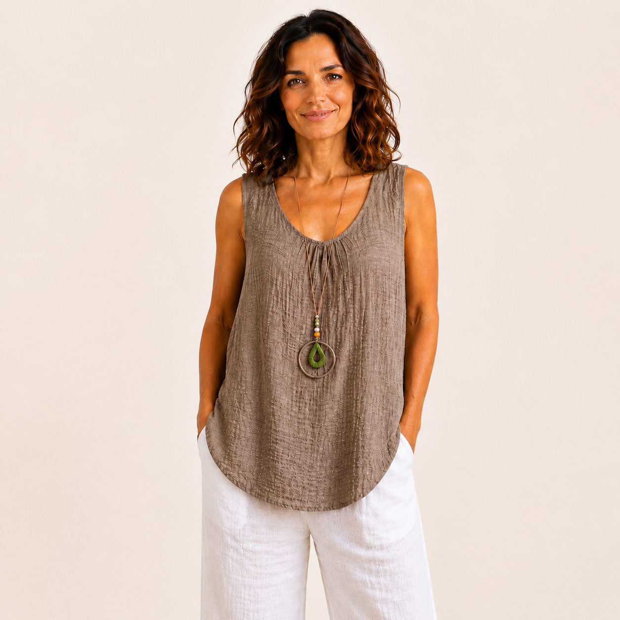 Woman modelling a relaxed-fit textured sleeveless tank top with a scoop neckline and curved hem, styled with white wide-leg linen pants and a long pendant necklace. Lightweight crinkle-textured fabric drapes comfortably over the body, shown in Black, Brown, Khaki, Light Blue, Pink, and White colour options. One-size style with a loose, flattering fit suitable up to a size 16.
