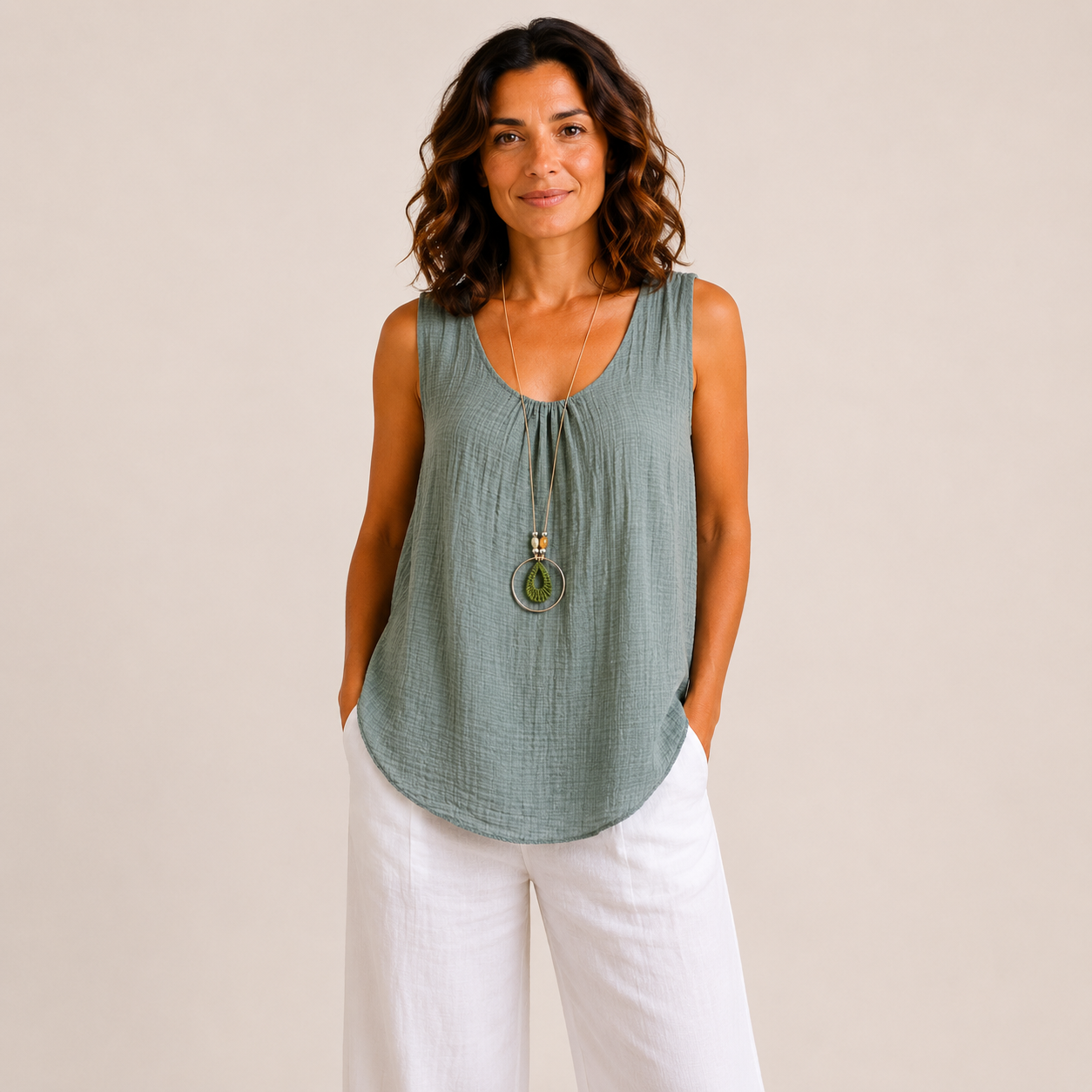 Woman modelling a relaxed-fit textured sleeveless tank top with a scoop neckline and curved hem, styled with white wide-leg linen pants and a long pendant necklace. Lightweight crinkle-textured fabric drapes comfortably over the body, shown in Black, Brown, Khaki, Light Blue, Pink, and White colour options. One-size style with a loose, flattering fit suitable up to a size 16.