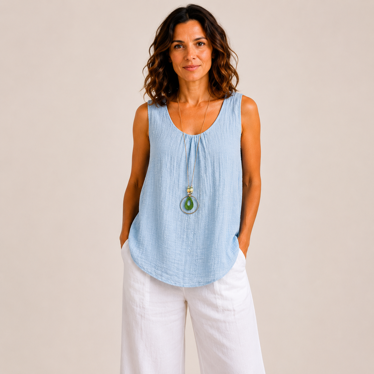 Woman modelling a relaxed-fit textured sleeveless tank top with a scoop neckline and curved hem, styled with white wide-leg linen pants and a long pendant necklace. Lightweight crinkle-textured fabric drapes comfortably over the body, shown in Black, Brown, Khaki, Light Blue, Pink, and White colour options. One-size style with a loose, flattering fit suitable up to a size 16.