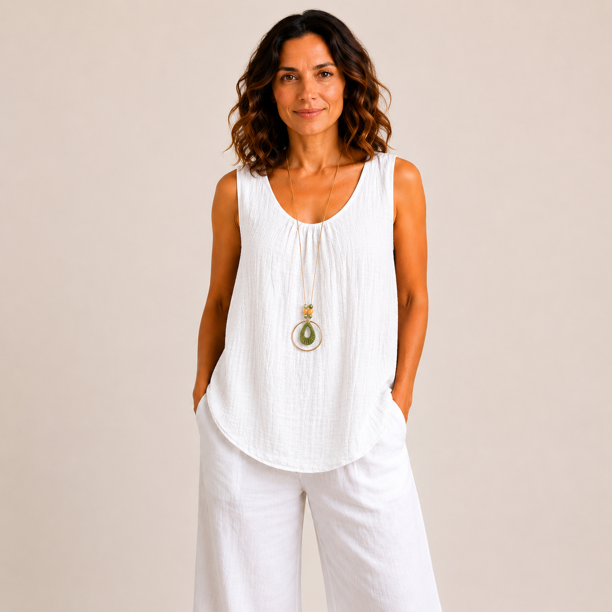 Woman modelling a relaxed-fit textured sleeveless tank top with a scoop neckline and curved hem, styled with white wide-leg linen pants and a long pendant necklace. Lightweight crinkle-textured fabric drapes comfortably over the body, shown in Black, Brown, Khaki, Light Blue, Pink, and White colour options. One-size style with a loose, flattering fit suitable up to a size 16.