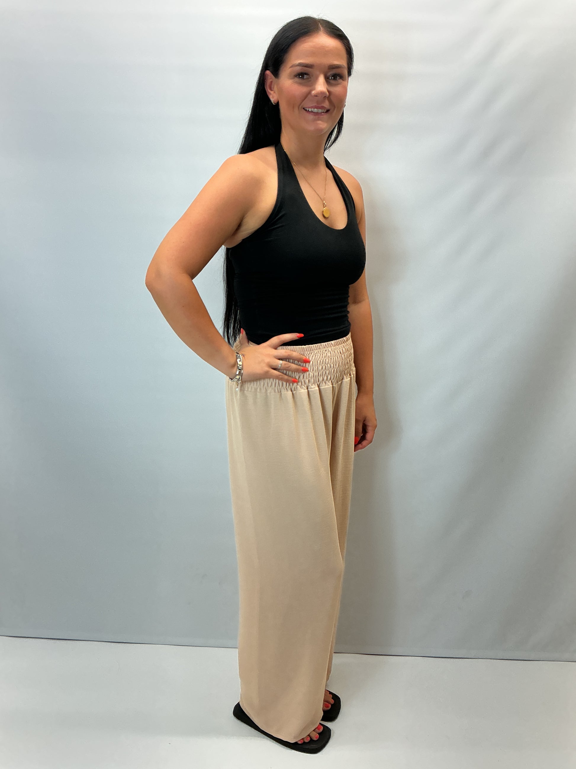 Beige smocked waist wide leg trousers with a relaxed fit, shown on a model wearing a fitted black halter top.