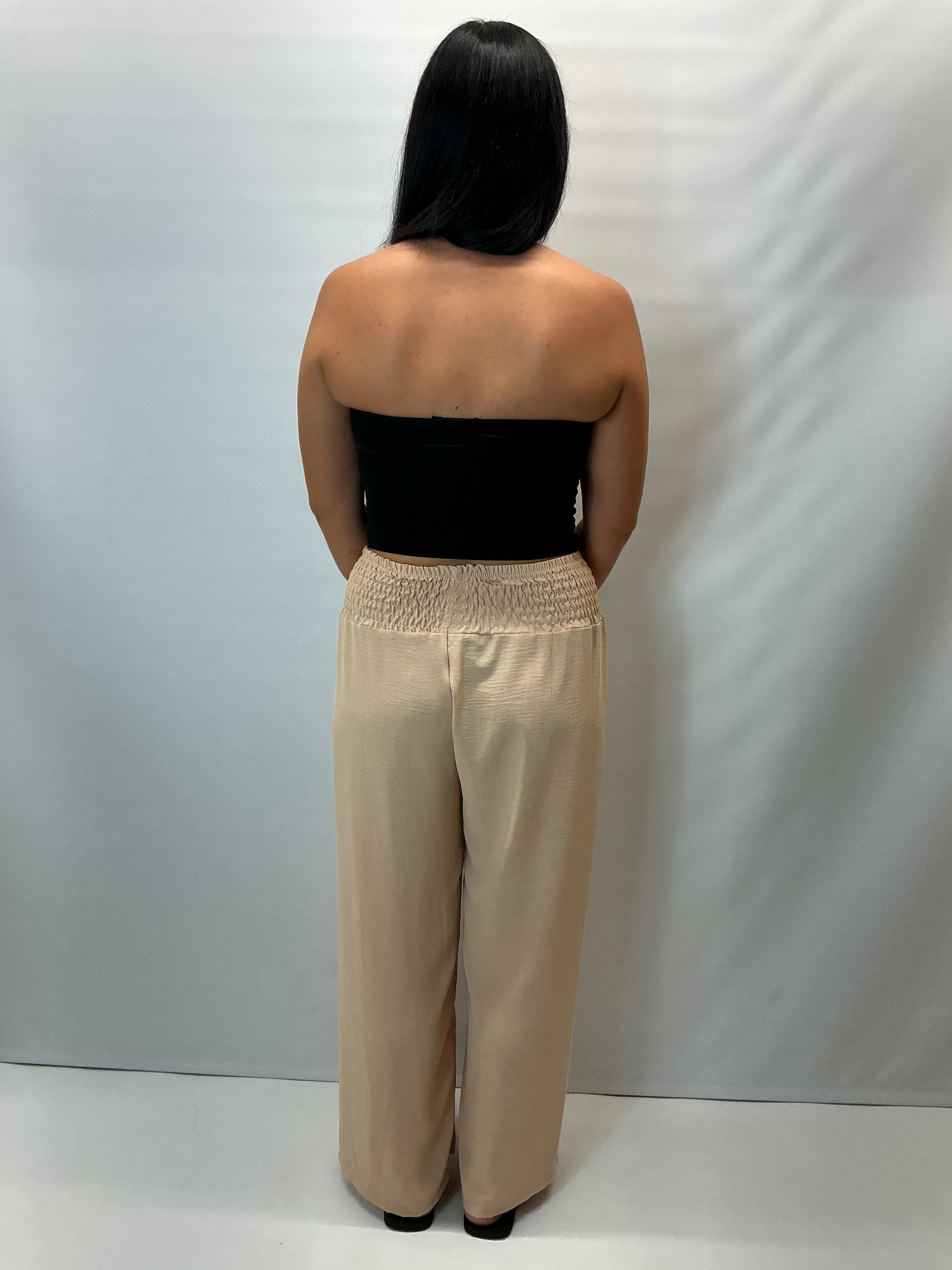 Beige smocked waist wide leg trousers with a relaxed fit, shown on a model wearing a fitted black halter top.