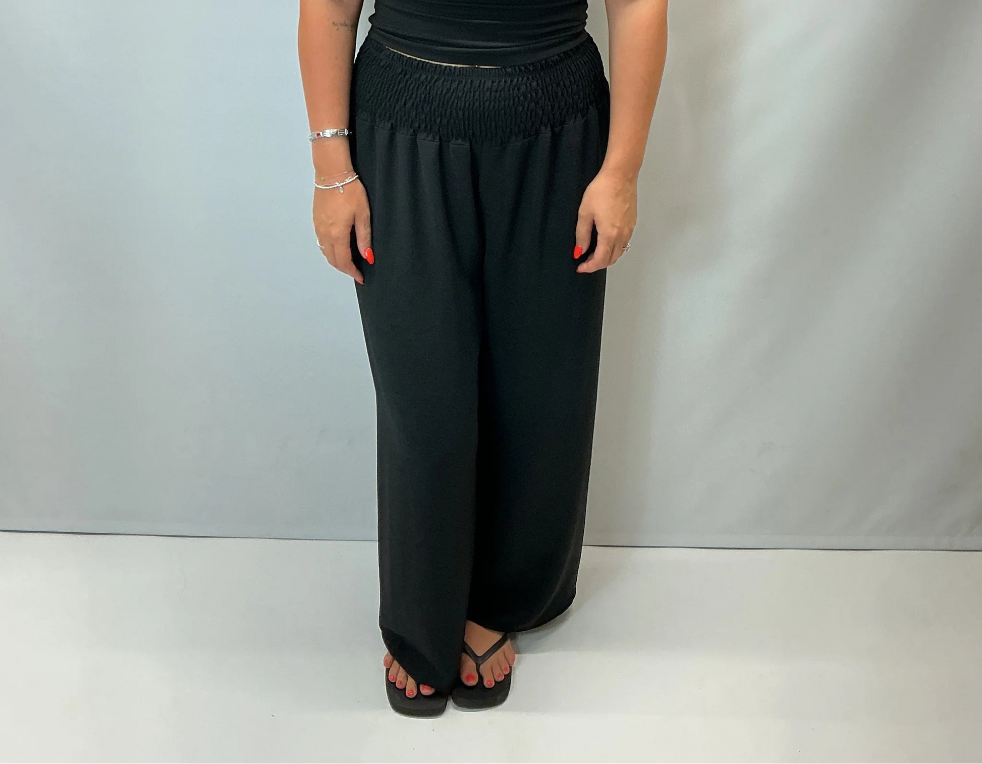 Black smocked waist wide leg trousers with a relaxed fit, shown on a model wearing a fitted black halter top.