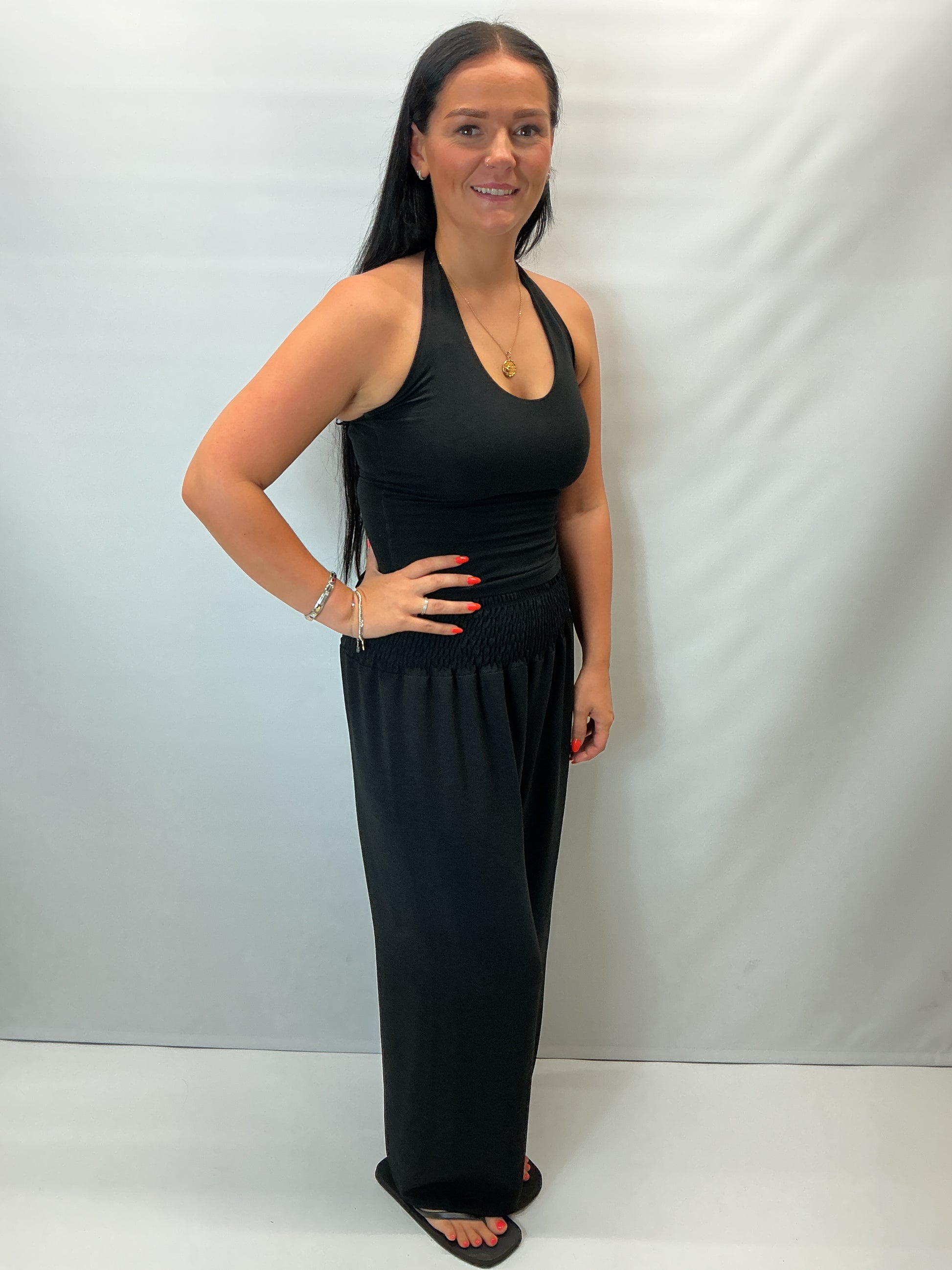 Black smocked waist wide leg trousers with a relaxed fit, shown on a model wearing a fitted black halter top.