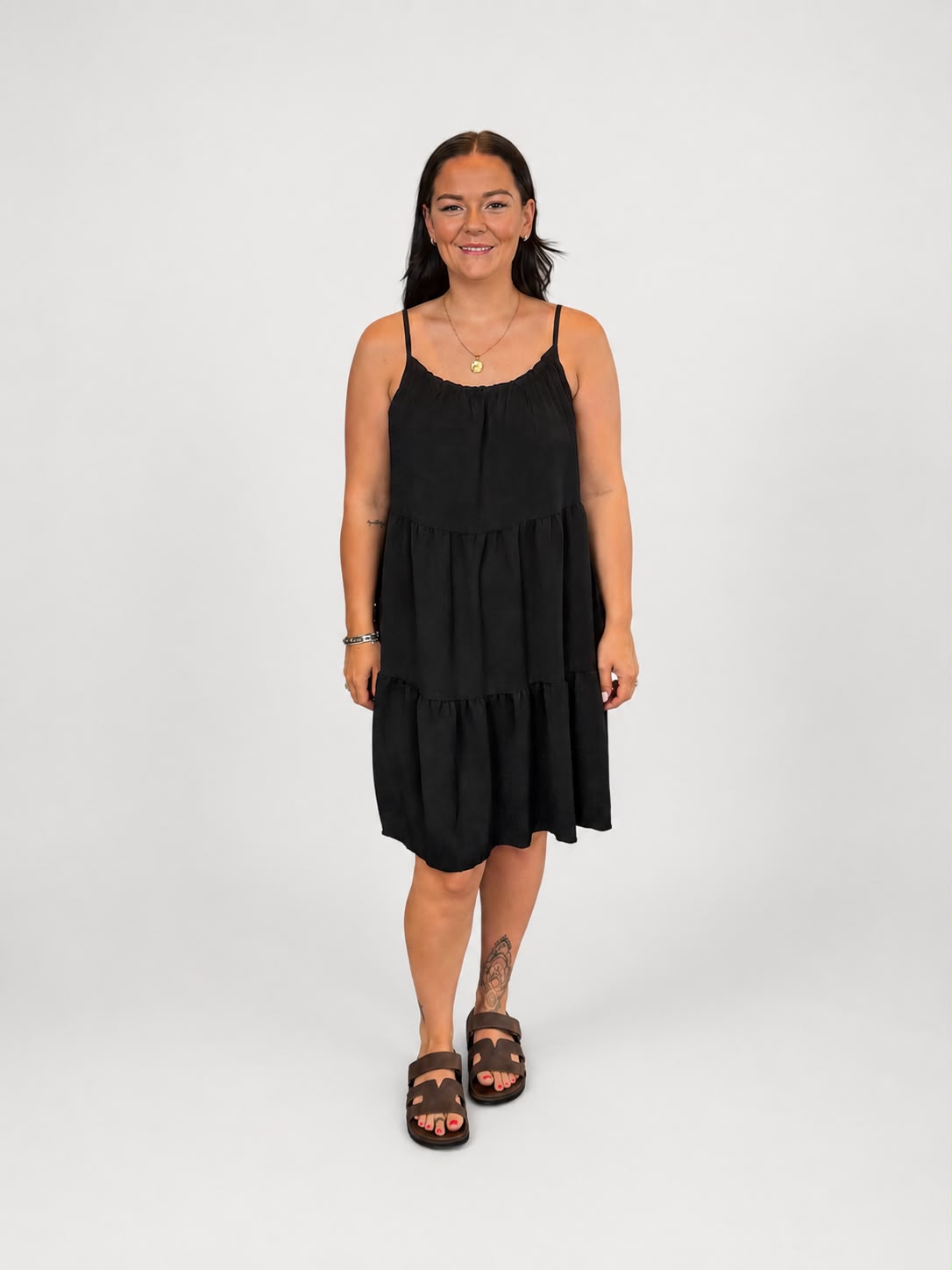 A versatile black tiered cami dress featuring spaghetti straps, a relaxed, flowy fit, and a flattering knee-length design. Lightweight and easy to wear, it's ideal for casual everyday styling, holidays, weekends, and warm-weather occasions.