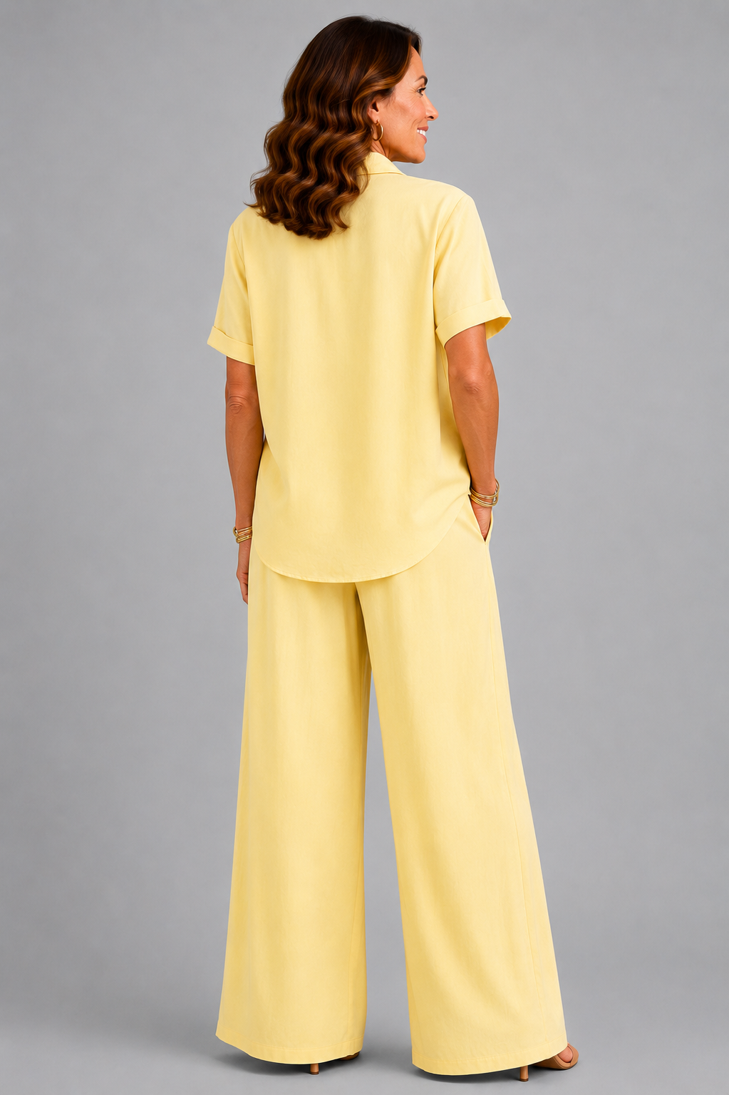 A smiling woman models a relaxed-fit Italian two-piece co-ord set on a light grey studio background. The outfit features a collared short-sleeve top with a detachable patterned scarf and matching wide-leg trousers. Shown in multiple colourways including black, cornflower blue, powder blue, sage green, blush pink and lemon yellow, the set offers an elegant, comfortable silhouette ideal for everyday wear and special occasions.