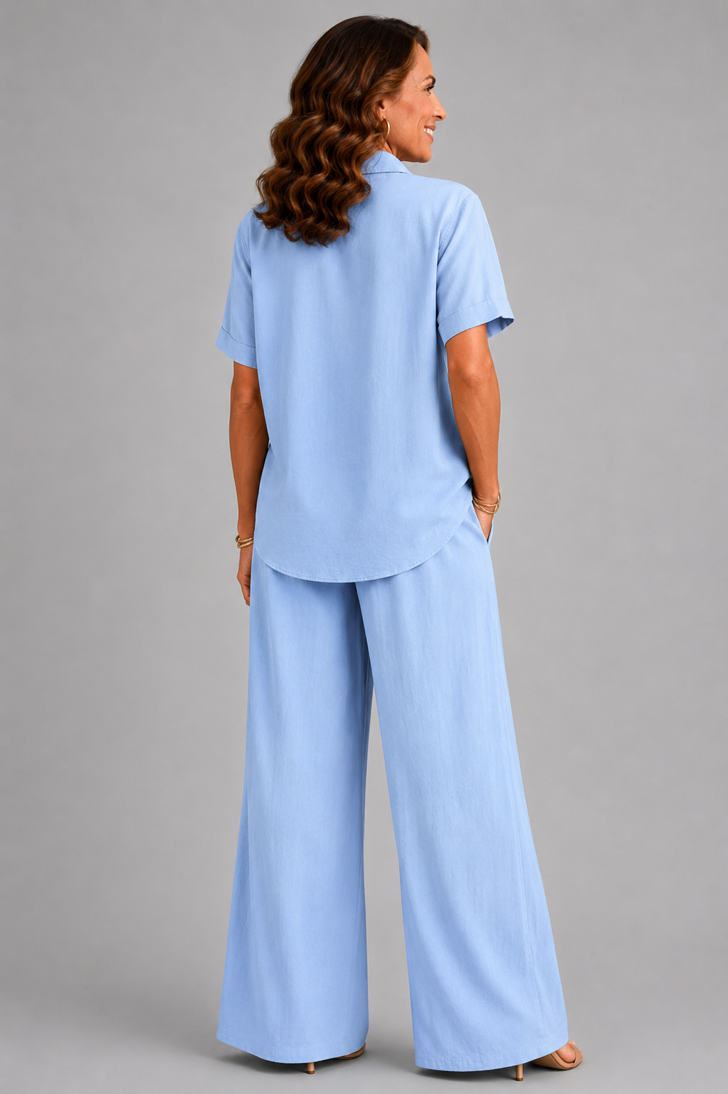 A smiling woman models a relaxed-fit Italian two-piece co-ord set on a light grey studio background. The outfit features a collared short-sleeve top with a detachable patterned scarf and matching wide-leg trousers. Shown in multiple colourways including black, cornflower blue, powder blue, sage green, blush pink and lemon yellow, the set offers an elegant, comfortable silhouette ideal for everyday wear and special occasions.