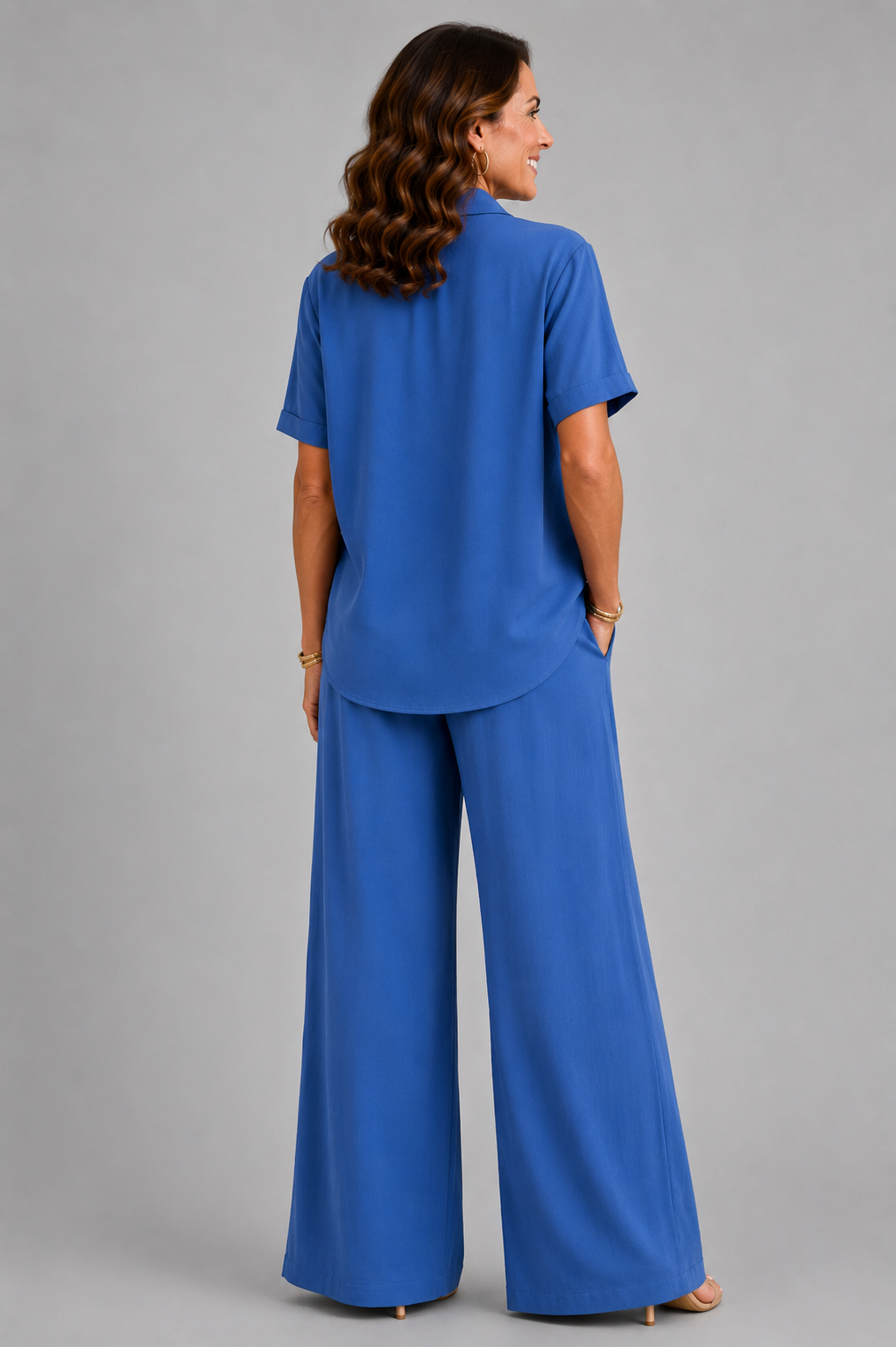 A smiling woman models a relaxed-fit Italian two-piece co-ord set on a light grey studio background. The outfit features a collared short-sleeve top with a detachable patterned scarf and matching wide-leg trousers. Shown in multiple colourways including black, cornflower blue, powder blue, sage green, blush pink and lemon yellow, the set offers an elegant, comfortable silhouette ideal for everyday wear and special occasions.