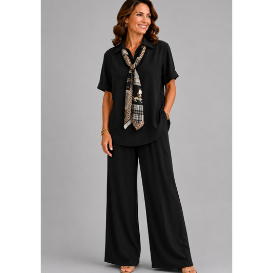 A smiling woman models a relaxed-fit Italian two-piece co-ord set on a light grey studio background. The outfit features a collared short-sleeve top with a detachable patterned scarf and matching wide-leg trousers. Shown in multiple colourways including black, cornflower blue, powder blue, sage green, blush pink and lemon yellow, the set offers an elegant, comfortable silhouette ideal for everyday wear and special occasions.