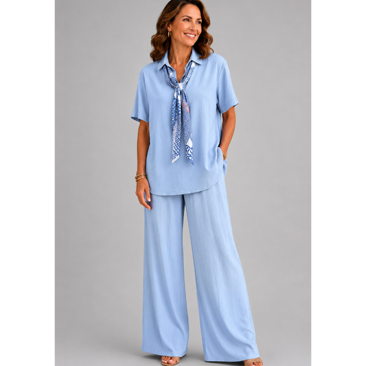 A smiling woman models a relaxed-fit Italian two-piece co-ord set on a light grey studio background. The outfit features a collared short-sleeve top with a detachable patterned scarf and matching wide-leg trousers. Shown in multiple colourways including black, cornflower blue, powder blue, sage green, blush pink and lemon yellow, the set offers an elegant, comfortable silhouette ideal for everyday wear and special occasions.