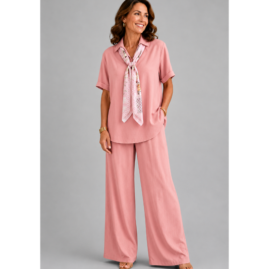 A smiling woman models a relaxed-fit Italian two-piece co-ord set on a light grey studio background. The outfit features a collared short-sleeve top with a detachable patterned scarf and matching wide-leg trousers. Shown in multiple colourways including black, cornflower blue, powder blue, sage green, blush pink and lemon yellow, the set offers an elegant, comfortable silhouette ideal for everyday wear and special occasions.