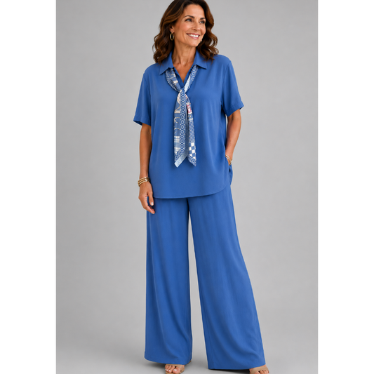 A smiling woman models a relaxed-fit Italian two-piece co-ord set on a light grey studio background. The outfit features a collared short-sleeve top with a detachable patterned scarf and matching wide-leg trousers. Shown in multiple colourways including black, cornflower blue, powder blue, sage green, blush pink and lemon yellow, the set offers an elegant, comfortable silhouette ideal for everyday wear and special occasions.