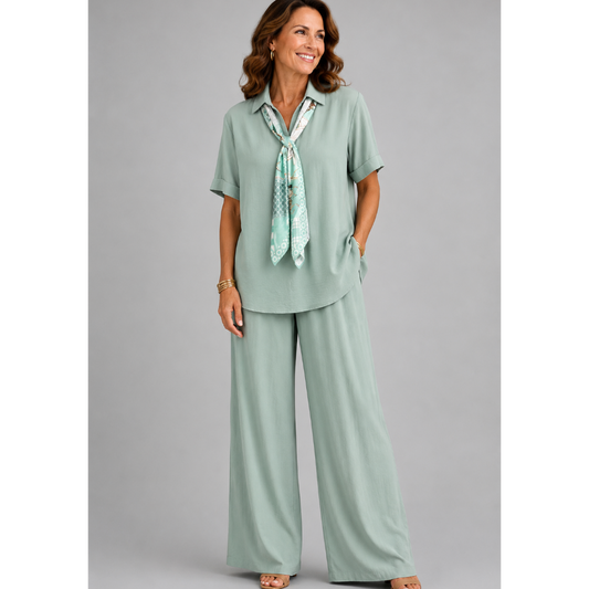 A smiling woman models a relaxed-fit Italian two-piece co-ord set on a light grey studio background. The outfit features a collared short-sleeve top with a detachable patterned scarf and matching wide-leg trousers. Shown in multiple colourways including black, cornflower blue, powder blue, sage green, blush pink and lemon yellow, the set offers an elegant, comfortable silhouette ideal for everyday wear and special occasions.
