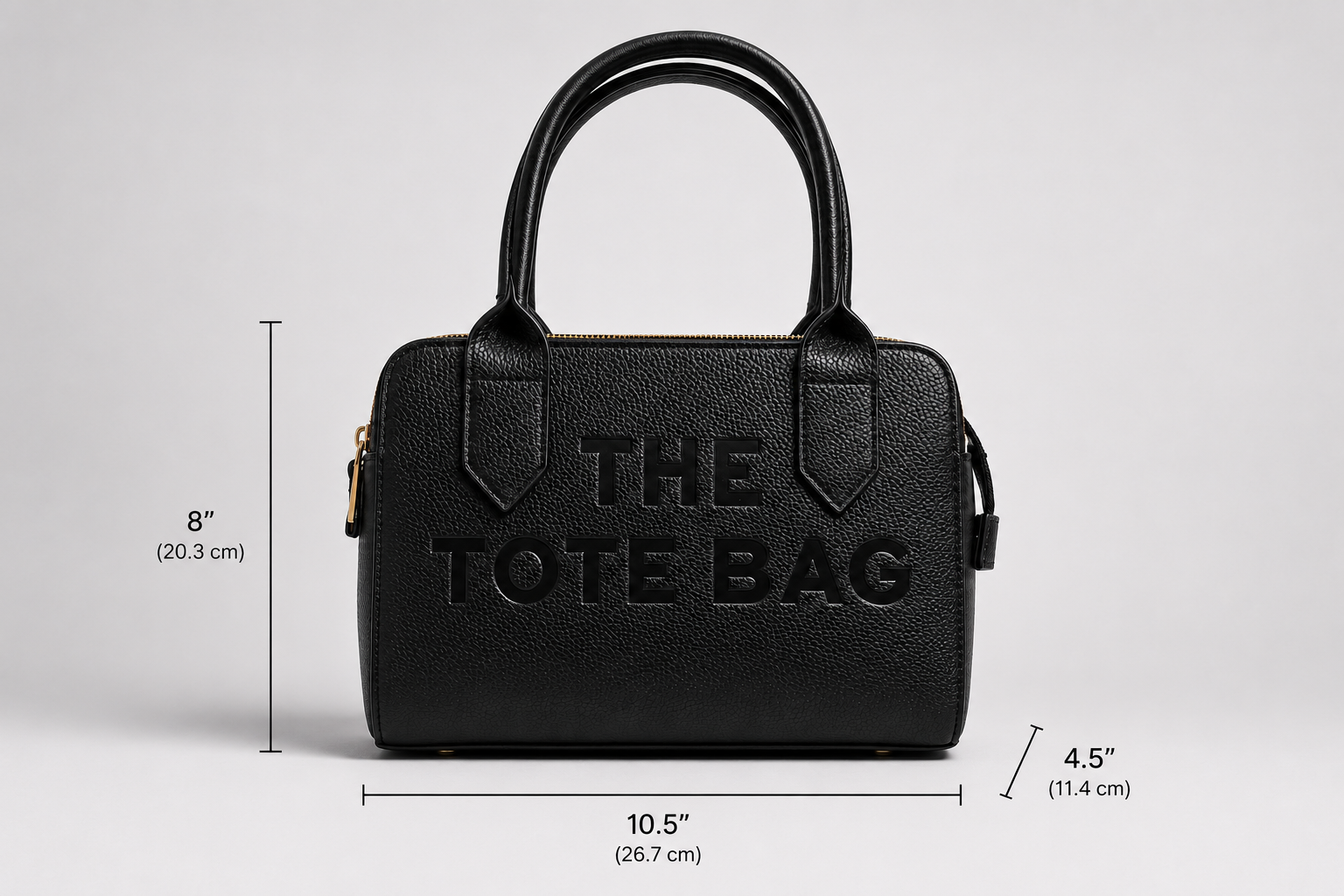 The Chelsea Tote Bag in Black featuring a structured design, pebbled faux leather finish, double zip compartments, gold-tone hardware, and embossed logo detail on a light grey studio background.