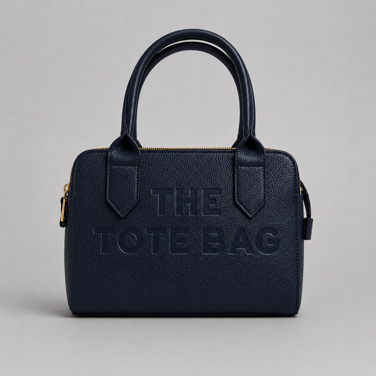 The Chelsea Tote Bag in Navy Blue featuring a structured design, pebbled faux leather finish, double zip compartments, gold-tone hardware, and embossed logo detail on a light grey studio background.