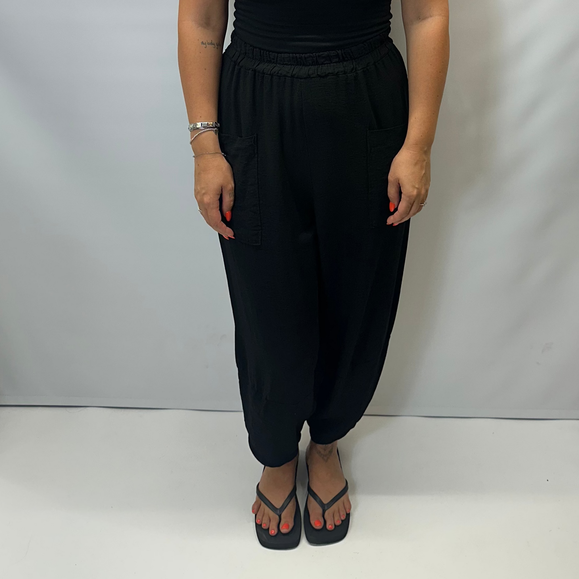 Model wearing black Italian relaxed tapered trousers with an elasticated waistband, front patch pockets and crossover split hem detail.